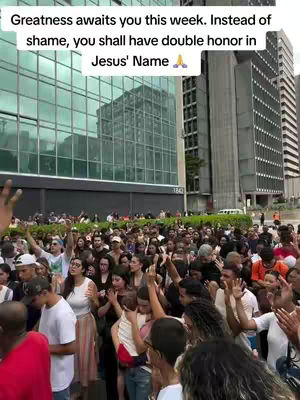 A large crowd of people is gathered outdoors, appearing to be at a rally or religious gathering. Many have their hands raised in the air. Skyscrapers and a large glass building are in the background with the text overlayed on the top portion of the image in a white box that reads: Greatness awaits you this week. Instead of shame, you shall have double honor in Jesus' Name with a praying hands emoji.