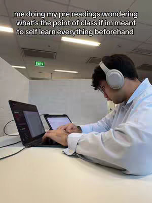 A person is sitting at a desk with two laptops in front of them, wearing headphones and glasses. The person is typing on one of the laptops, which has content displayed on its screen. On the wall is a green exit sign. Text overlayed onto the image reads: "me doing my pre readings wondering what's the point of class if im meant to self learn everything beforehand".