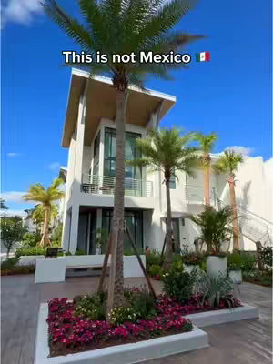 A modern, multi-story white building with large glass windows and a flat roof is shown. Palm trees and other greenery surround the building, including flowering plants in the foreground. The sky is bright blue. Text overlay at the top says "This is not Mexico".