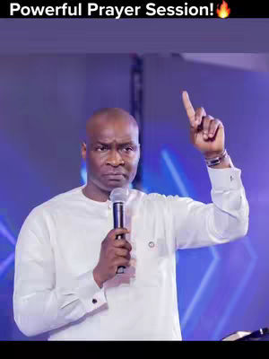 A man in a white shirt is gesturing with his right index finger raised towards the ceiling. He holds a microphone in his left hand. The background is a blurry mix of blue and purple hues.