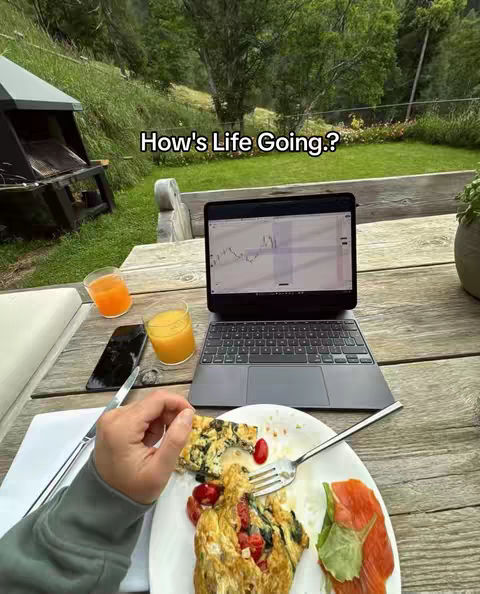 A person is seated outside at a wooden table, partaking in a casual breakfast or brunch. The table is set with a plate holding a portion of food, a laptop displaying what appears to be a trading chart, a glass of orange juice, a mobile phone etc. The background features a grassy area with trees, suggesting an outdoor setting. The lighting is natural, giving the scene a relaxed feel