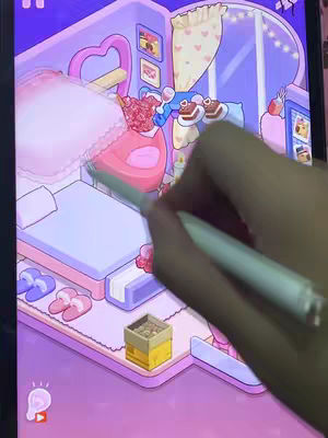 A screenshot from an app showing a digitally rendered, pastel-colored bedroom interior. A hand holding a stylus hovers over the screen, seemingly interacting with the app. The room features a bed, a vanity area with a heart-shaped mirror, and various decorative elements, with an overall aesthetic of a dream bedroom.
