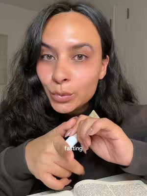 A medium shot of a woman with dark hair, looking slightly to the side with a contemplative expression. The text 'fasting' is overlaid on her hands. She is holding a small white object, possibly a pen or a container.