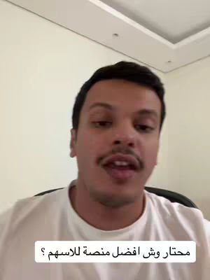 A medium shot shows a man in a white t-shirt speaking directly to the camera. The background is a plain white wall with a square ceiling detail. Text in Arabic appears at the bottom of the screen.