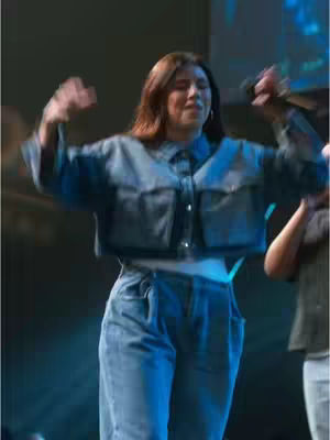 A medium shot of a woman on stage, likely at a concert or church service. She is wearing a denim jacket and matching or coordinating pants, with her arms raised in a gesture of praise or excitement. The background is blurred but appears to be a stage with lights and possibly other performers.