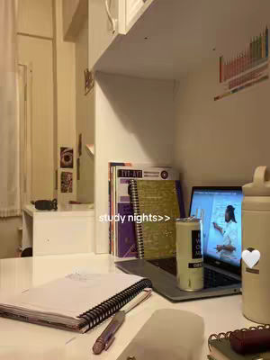 The image shows a well-lit desk with study materials. A laptop displays a video of a person. Visible are books, a notebook, a pen, and a beverage can. The background is somewhat minimalistic, with storage shelves and a window.