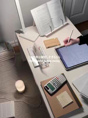The image shows a well-lit desk setup, with a book open on a stand, a tablet, a calculator, and other study materials. A hand is holding a stylus, and the overall ambiance is calm and focused.