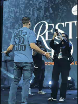 The image shows a live event setting, likely a church conference. The focus is on three individuals, one wearing a blue t-shirt with a prominent 'GO TO GOD' graphic, turned toward the camera. Behind them, a large banner reads 'REST', with other lettering obscured by the individuals. The lighting indicates a stage setting, with a soft backdrop and visual elements of a live presentation. The image captures an active moment, showing people engaged in the event.