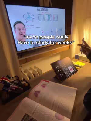 A well-lit indoor scene with a desk and a large TV screen displaying a medical diagram. A man's face is superimposed on the TV screen. Study materials, a tablet, and pens are scattered on the desk in the foreground.