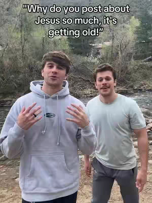 Two young men stand outdoors with a natural backdrop. The man on the left wears a light gray hoodie and gestures with open hands, while the man on the right is in a light green t-shirt, his arms at his sides. The background shows trees and a flowing body of water.