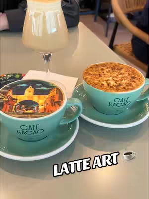 The image showcases three different lattes presented on a gray surface.  The first latte, in a tall glass, has a light beige hue and a foamy top.  The second and third lattes are in teal mugs with saucers, and feature a textured, golden-brown top, with the words 'CAFE KACAO' printed on it. The latte on the left has a coffee art design, a landscape with architectural elements. A small black coffee cup with a coffee art design appears towards the bottom right. Written in a bold, white font across the bottom is the phrase ' LATTE ART'.  The scene is well-lit, casting soft shadows and suggesting an inviting brunch atmosphere.