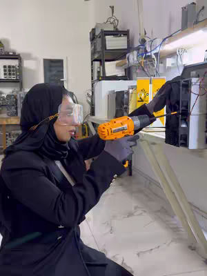 A woman wearing a black headscarf, safety glasses, and gloves uses an orange drill on a black piece of equipment in what appears to be a workshop or lab setting. The background includes white and yellow equipment, wires, and shelving.