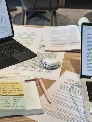 The image shows a close-up of a desk with multiple scattered elements suggesting an active study or work session. There are two laptops, one on the left with a keyboard and another on the right displaying a screen filled with text. Several printed pages are spread across the desk, some filled with text and others with handwritten notes. A pink pen and a pair of white AirPods with charging case add to the workspace setting. Two sticky notes with handwritten notes are on the left of the image. The setup creates a sense of productivity, focused work, along with the study-related notes around.