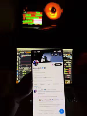 A person's hand holds a smartphone displaying a social media profile. The background includes a computer monitor with trading charts and a glowing orange light. The image is generally dark with pops of color from the screens.