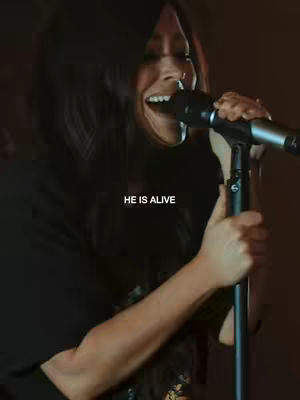 A woman singing into a microphone, her mouth open in song and eyes closed. The text 'HE IS ALIVE' in white is superimposed on the image.