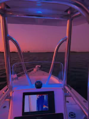 The image is taken from inside a boat, looking towards the horizon. The foreground features the boat's metallic frame and dashboard, with a screen displaying what appears to be sonar data. The color palette is dominated by shades of purple and orange, suggesting a sunset or sunrise.
