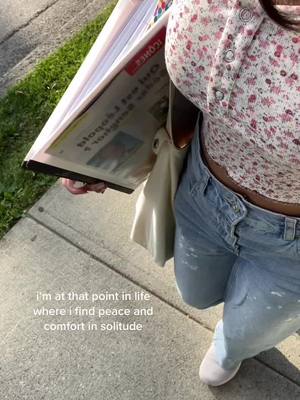 The image is a high-angle shot of a person's midsection while walking outside, with the camera pointing down. The person is wearing a button-down floral top with a soft palette of light pink and white. They are sporting light-wash, distressed jeans and light-colored sneakers. They are carrying a large, open folder with the newspaper partially visible.