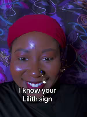 A close-up of a smiling woman with dark skin wearing a red head covering and a black top. She has elaborate nose jewelry, visible make-up, and her mouth is open, revealing a red lipstick. The background is a stylized patterned fabric. Text at the bottom reads, "I know your Lilith sign."