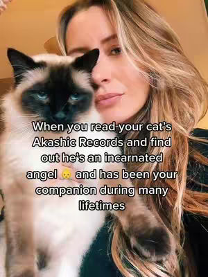 A woman with long blonde hair holds a Siamese cat close to her face. The cat has distinctive dark markings on its face and ears. The woman is looking at the camera with a slight, inward expression, potentially suggesting introspection or a shared moment with the cat. Overlayed text provides the context of the video.