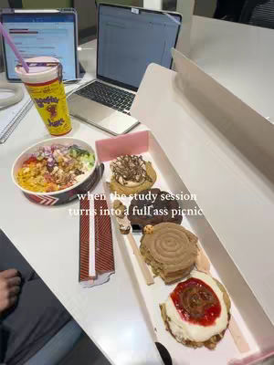 A top-down shot of a white table overflowing with food: a bowl of colorful food, a drink, a laptop, and a box of cookies. The text overlayed says "when the study session turns into a full ass picnic".