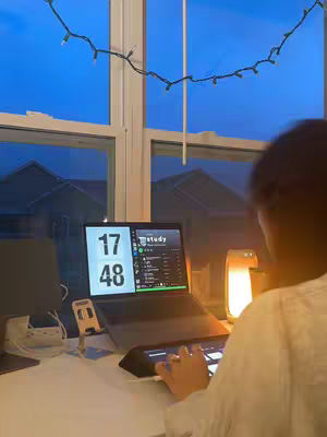 The image shows a person from the back sitting at a desk with a laptop open. Behind the person is a window with a view of houses and a blue sky, with a string of lights hanging in front. The laptop screen is split showing a digital clock and what appears to be a music app.