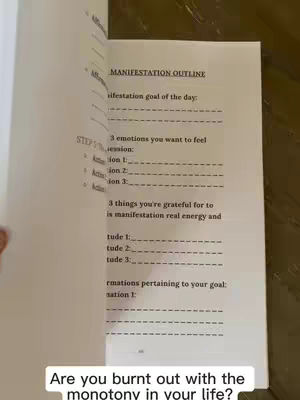 An open book displaying a worksheet titled "MANIFESTATION OUTLINE". The pages are white with black text and horizontal lines for writing prompts. The book lies on a brown surface, and a hand is visible, presumably turning the pages.