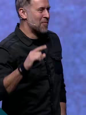 A man in a black button-down shirt is gesturing with his right index finger, appearing to make a point or emphasize a statement. The background is a blurry blue.