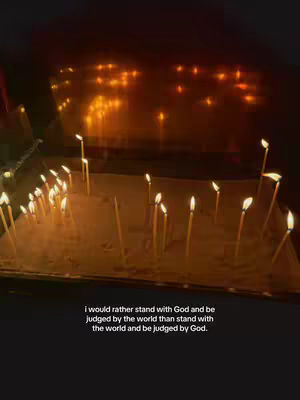 A dimly lit interior shot focuses on numerous lit candles arranged on a sandy surface. The candles cast a warm, flickering light, and their flames are prominent. Below, a text overlay provides a quote.