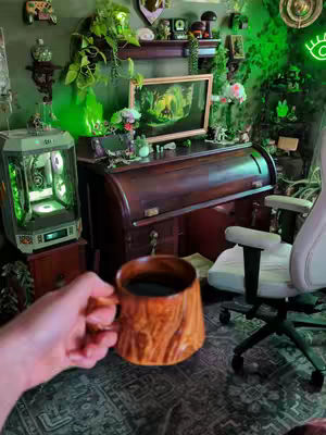 The scene shows a cozy gaming setup with a wooden rolltop desk, a gaming PC, and various decorations creating a maximalist aesthetic. The focus is on a mug held by a hand, suggesting the creator is enjoying the space.