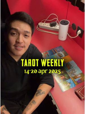 A man with dark hair, wearing a black hoodie, sits at a red desk with tarot cards spread out. The background is a red wall with a power outlet. Text overlayed on the image says 'TAROT WEEKLY' above '14-20 apr 2025'.