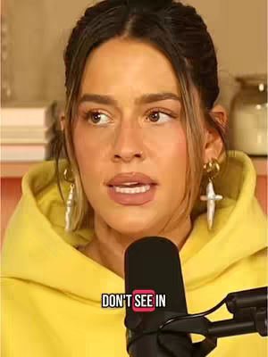 The image features a woman with light skin, brown eyes, and medium-length brown hair with blonde highlights. She's wearing a yellow hoodie and large, gold earrings in the shape of crosses. A microphone is positioned in front of her, and a text overlay reads "DON'T SEE IN". The background is blurred, suggesting an indoor setting.
