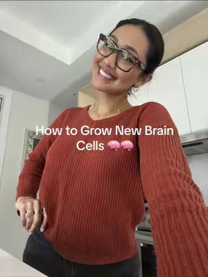 A woman in a red sweater is in a well-lit kitchen, smiling at the camera while wearing glasses. Text overlayed on the image reads, "How to Grow New Brain Cells," accompanied by brain emojis. The background includes kitchen cabinets and appliances.