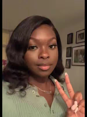 A woman with dark hair and a green shirt is holding up two fingers in a peace and love gesture. She is wearing a necklace with the word 'Grace' on it. The background shows a wall with framed pictures.