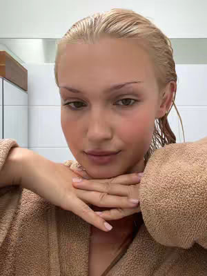 The image shows a close-up of a person with a light complexion, likely a woman, in a bathroom setting. Her wet, blonde hair is slicked back. She's wearing a brown bathrobe. Her hands are folded under her chin. The background consists of a white wall, a mirror, and a wooden cabinet. The lighting is soft and natural.