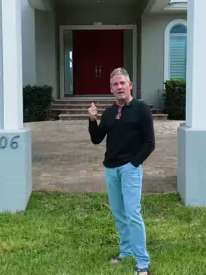 A man in a black shirt and blue jeans stands in front of a large house. He is gesturing with a thumbs-up, and a house is visible in the background.
