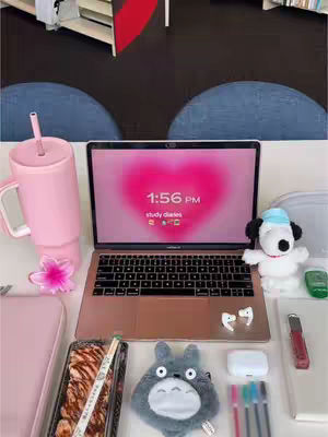 A top-down view of a desk setup for studying. The desktop features a pink laptop displaying a heart-shaped gradient with the time '1:56 PM'. Various study-related items surround the laptop, including a large pink water bottle, a plush Totoro, a Snoopy plush, and a tray of food. The background includes blue chairs, and a hint of a bookshelf.