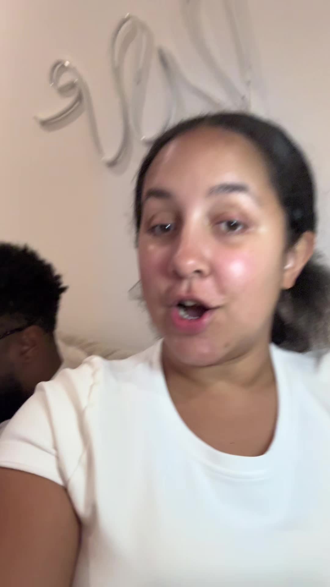 A selfie-style video shows a woman in the foreground with her mouth open, possibly speaking. Behind her, a man's blurred face and a portion of a wall with decorative elements are visible.