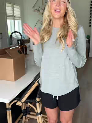 A woman with long blonde hair stands in a bright kitchen, wearing a light green long-sleeved shirt and black shorts. She gestures with both hands, palms up. A partially visible cardboard box sits on the counter to her left.