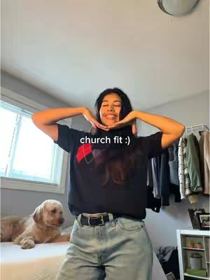 A young woman with long, dark hair, wearing a black t-shirt and light-wash jeans, stands in a well-lit bedroom. She has her arms up, framing her face with her hands. Behind her, a dog is lying on a bed to the left, a window and some clothes hanging are visible. The text 'church fit :)' appears overlayed.