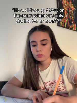 A young woman with long brown hair sits at a desk, looking pensively towards the viewer. She holds a pen and faces a piece of paper on the desk. The background includes a wall with a painting, creating a casual and studious atmosphere. A text overlay appears at the top, over her head.