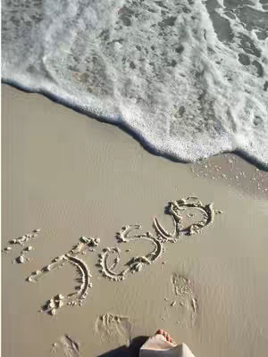 A photograph of a beach scene with the word "JESUS" written in the sand, with the ocean waves gently washing in. In the lower right, a person's feet are visible stepping on the sand.