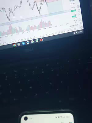 The image shows a laptop with a trading platform open, displaying a stock chart. Below the laptop is a phone. The composition is from a slightly high angle, focusing on the devices.