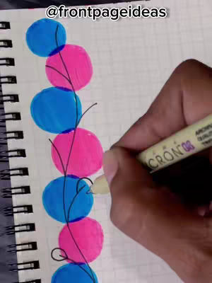 A hand is drawing a colorful design on a notebook page. The design consists of alternating blue and pink circles connected by thin black lines, forming a vertical pattern. The hand holds a cream-colored pen.