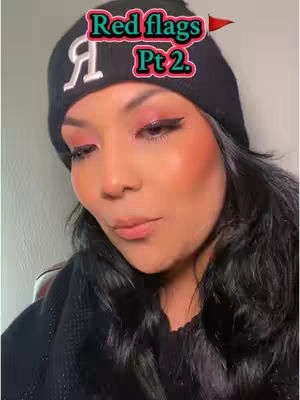 A close-up shot of a woman with dark hair, wearing a black beanie and a black scarf. The text overlay is a pink banner with text on her beanie. She has pink eye shadow and is looking slightly downward.