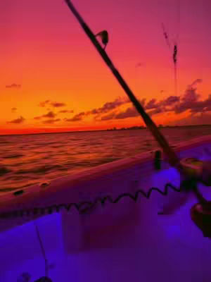 A vivid sunset scene over the ocean with a fishing rod and the side of a boat in the foreground. The sky transitions from orange to pink, clouds are visible towards the top of the frame. The boat features purple LED lighting.