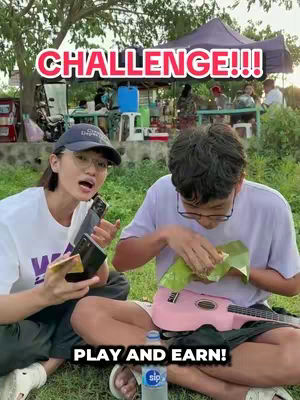 The cover image shows two people, a woman and a man, sitting on the grass outside. The woman, on the left, is holding two phones and looking towards the camera with her mouth open like she is talking. The man, on the right, is eating from the leaf-wrapped food and holding a pink ukulele. Text overlays say "CHALLENGE!!!" and "PLAY AND EARN!". A bottle of water sits at the bottom.