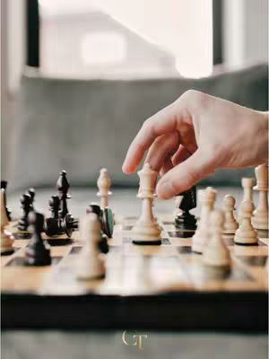 A hand moves a white queen chess piece on a chessboard. The board is wooden with black and white pieces, and the background is blurred with a window and a gray surface.