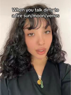 A woman with dark curly hair looks directly at the camera. Overlay text reads "When you talk dirty to a fire sun/moon/venus". She wears a black top with a pendant.