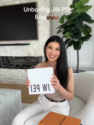 A woman with long black hair sits smiling in a well-lit living room. She holds a white sign that reads "139 WL", wearing a casual outfit. In the background, a large television is mounted, and a large houseplant sits next to her. There is a box in the front and a Louis Vuitton Box