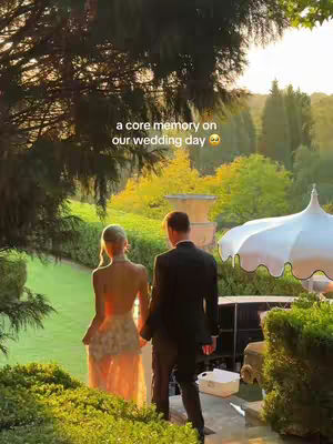 A couple, dressed in wedding attire, walks away from the camera towards a vibrant sunset. The bride wears a white or light-colored gown, while the groom is in a black suit. The scene is framed with trees and greenery, giving it a natural, romantic backdrop. There is text overlay at the top, and the scene is well lit and evokes a sense of intimacy and celebration.